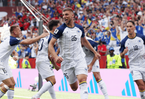 Soccer players in white and navy celebrate a goal on the field as the crowd cheers in the background.