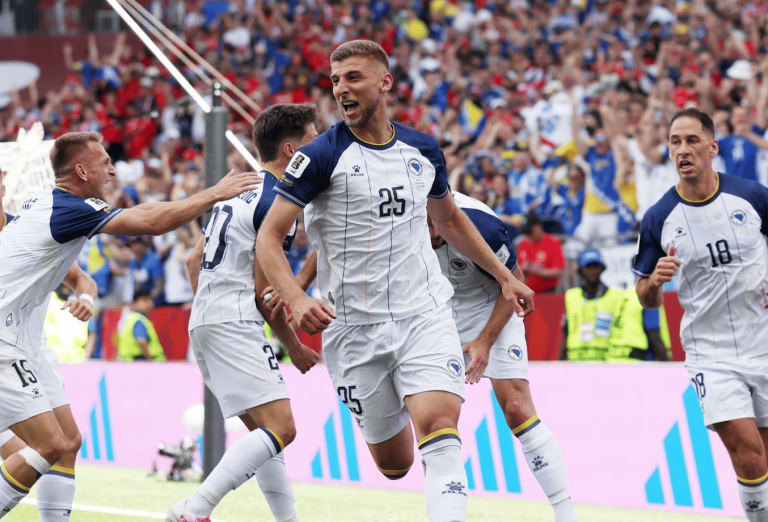 Soccer players in white and navy celebrate a goal on the field as the crowd cheers in the background.