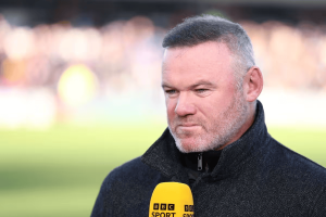 Man in a dark coat being interviewed on camera, speaking into a BBC Sport yellow microphone with a blurred stadium background