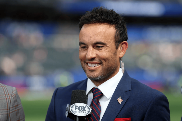 Smiling man in a navy suit and striped tie being interviewed at a sports event, with a FOX Sports microphone visible