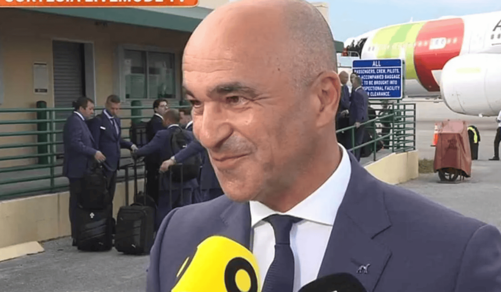 hupu-football-1781323956171-ayx-1png - AYX Sports Hub Man in a navy suit being interviewed by a yellow microphone at an airport, with colleagues and luggage in the background and a colorful airplane behind them.