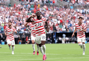 USA soccer players in red-and-white striped jerseys celebrate on a green pitch as a packed stadium cheers.