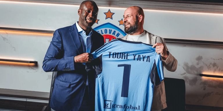 Two men pose in a modern room as one presents a light-blue soccer jersey reading 'TOURE YAYA' with the number 1.