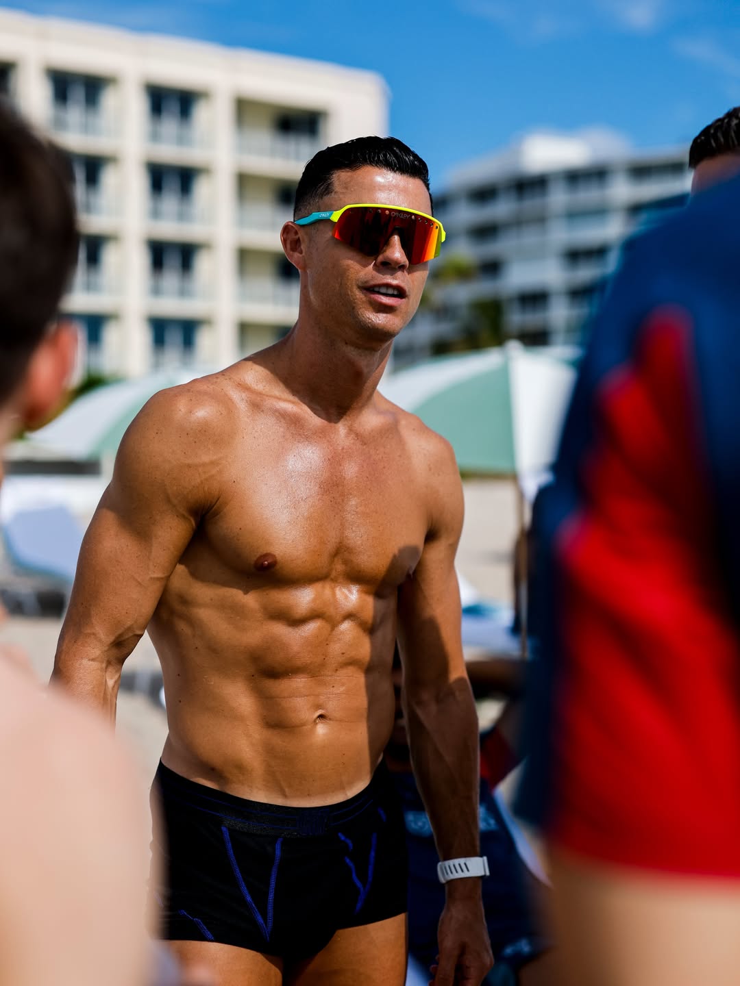 hupu-football-1781381478112-ayx-recoveryjpg - AYX Sports Hub Muscular, shirtless man wearing bright neon sunglasses stands on a sunny beach with resort buildings in the background.