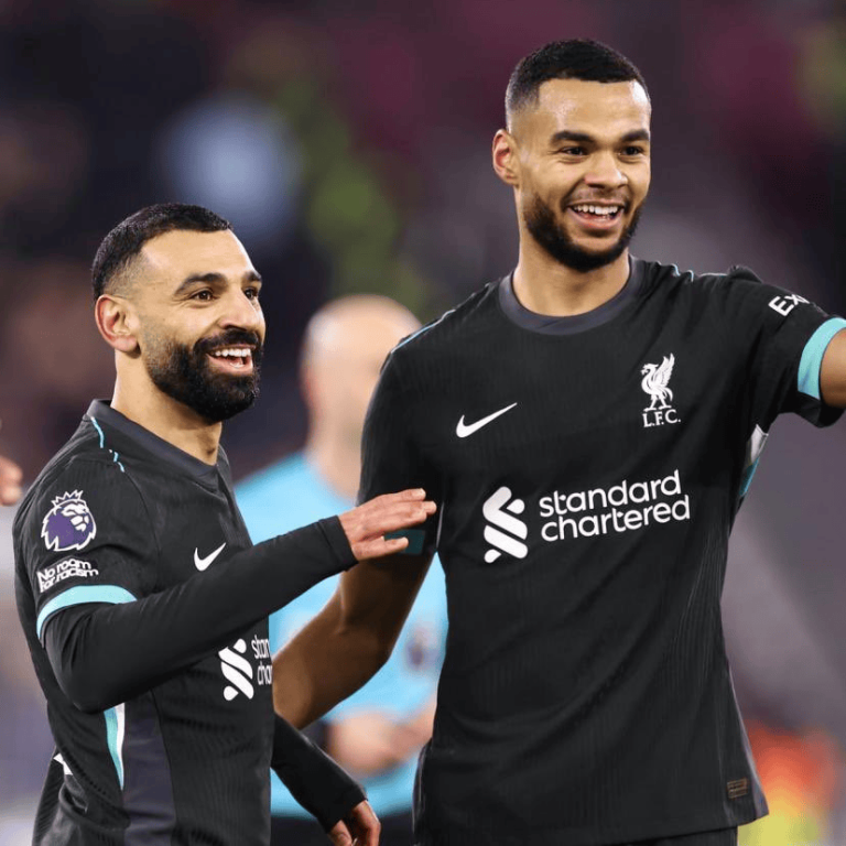 Two Liverpool FC players celebrate on the field, smiling in black away kits with sponsor logos visible on the front.