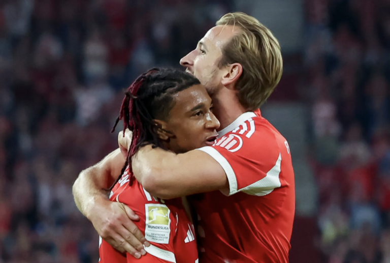 Two soccer players in red jerseys hug in celebration on the field, Bundesliga patch visible on one sleeve, stadium crowd in the background.