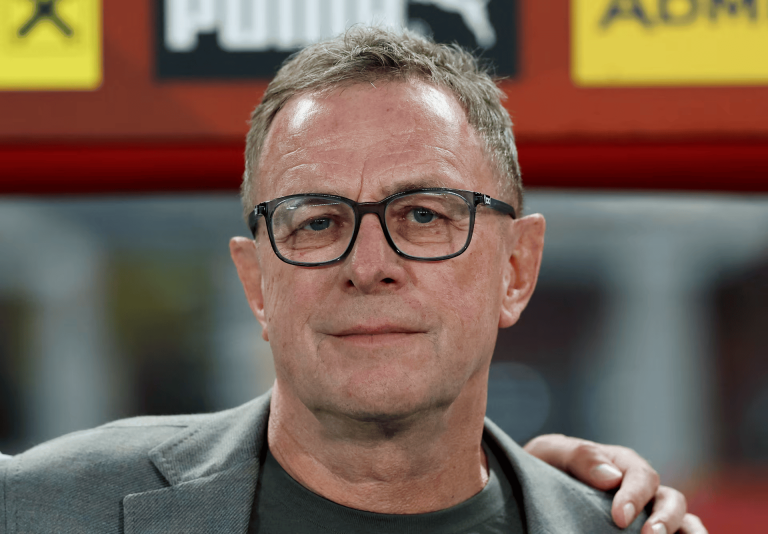 Close-up portrait of a middle-aged man with short gray hair and black-rimmed glasses, wearing a gray blazer and dark shirt, with a hand resting on his shoulder in the background.