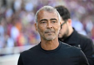 Portrait of a middle-aged man with gray hair and stubble wearing a black shirt, at a crowded stadium with blurred spectators behind him.
