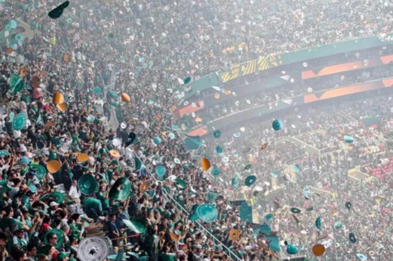 Crowd in a large stadium cheering as green and orange confetti and hats rain down during a celebration.