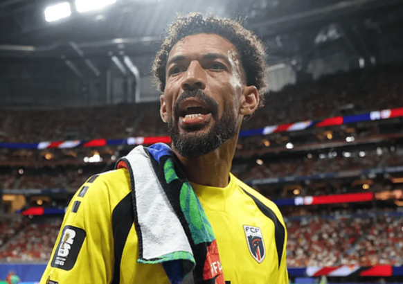 Male footballer in a bright yellow jersey with a towel over his shoulder, standing on a crowded stadium field after a match.