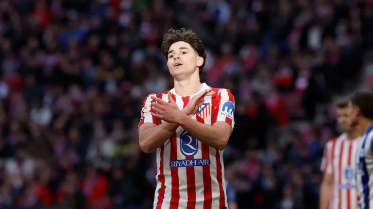 Soccer player in red-and-white striped jersey crossing arms in front of chest, celebrating on the field with blurred crowd behind him (Atletico Madrid, Riyadh Air sponsor).