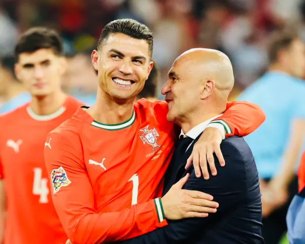 Portugal national team soccer player in a red jersey hugs a coach in a suit, both smiling on the field after a match.