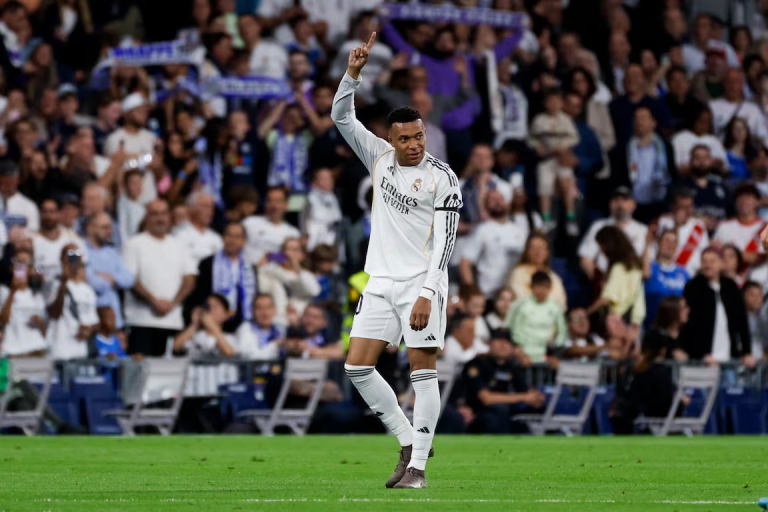 Soccer player in white Real Madrid kit raises one finger in celebration on a green pitch with cheering fans in the background.
