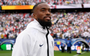 Male athlete with a beard wearing a white zip-up jacket, standing on a football field with a crowded stadium in the background.