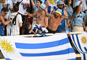 Crowd of soccer fans in blue and white, two shirtless men cheering behind a railing, Uruguay flags draped over the barrier.