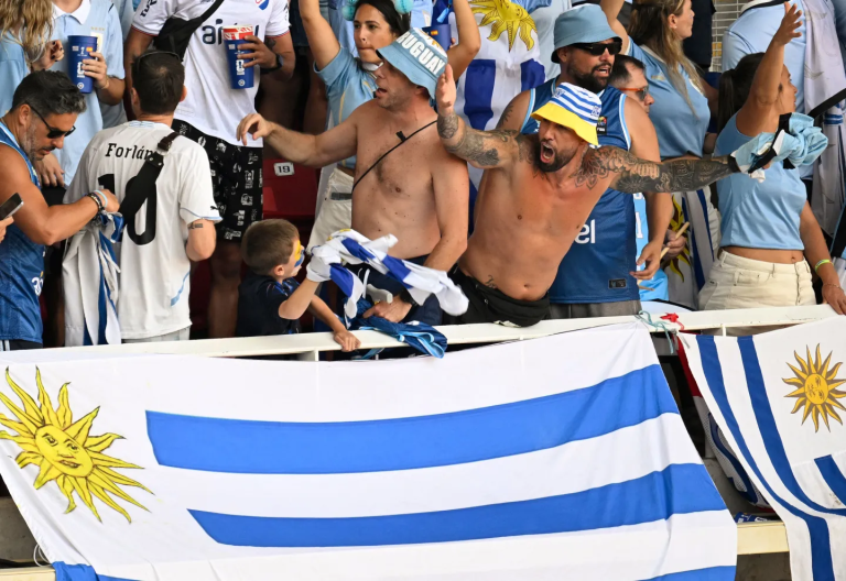 Crowd of soccer fans in blue and white, two shirtless men cheering behind a railing, Uruguay flags draped over the barrier.