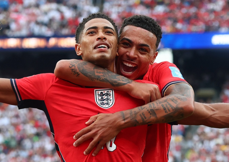 Two England footballers celebrate a goal with a joyful hug on the pitch as fans cheer in the background and wear red jerseys.