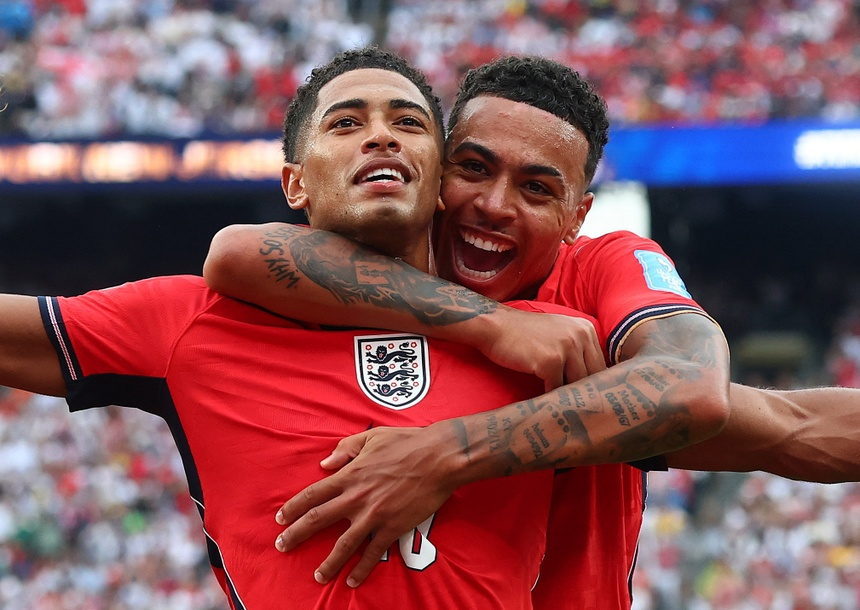 Two England footballers celebrate a goal with a joyful hug on the pitch as fans cheer in the background and wear red jerseys.