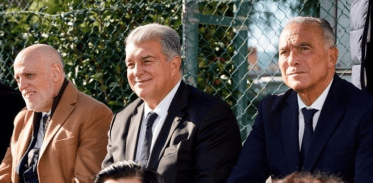 Three men in suits seated outdoors beside a chain-link fence, smiling and watching something.