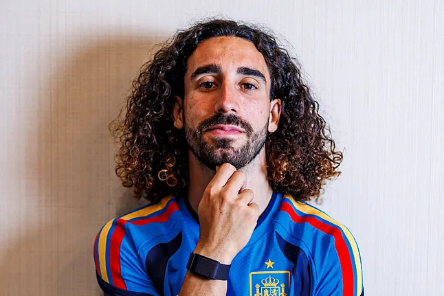 Portrait of a man with long curly hair and a beard, wearing a blue Spain jersey, resting his chin on his hand against a light wall.