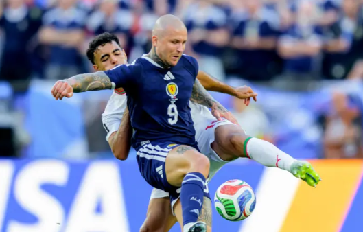 Soccer player in a blue kit (number 9) challenges an opponent for the ball during a match on a crowded stadium field.