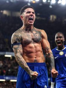 Tattooed footballer shirtless in blue shorts celebrating with clenched fists on a stadium pitch.