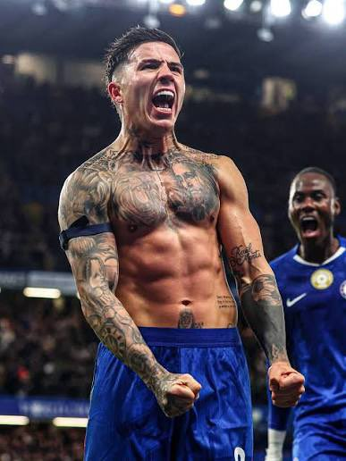 Tattooed footballer shirtless in blue shorts celebrating with clenched fists on a stadium pitch.