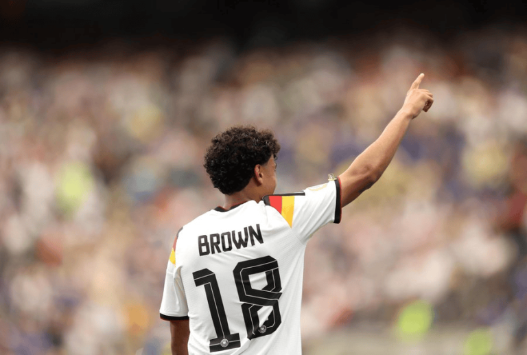 Soccer player in a white jersey with the name BROWN and number 18, raising his right arm in celebration.