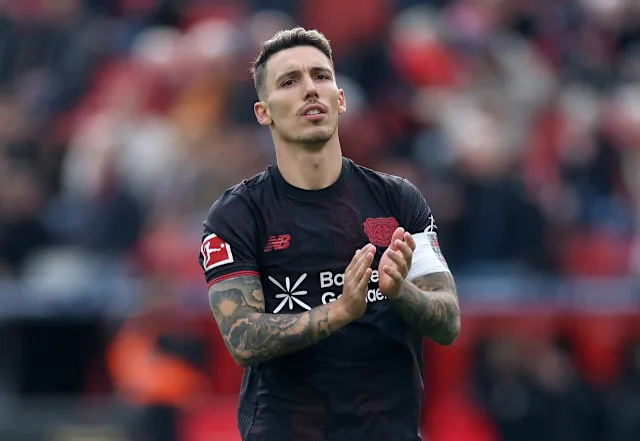 Soccer player in a black away kit clapping on the field with a blurred crowd in the background and tattooed arms.