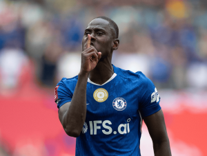 Soccer player in a blue Chelsea jersey making a shushing gesture with a finger to lips.