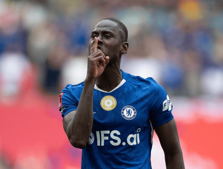 Soccer player in a blue Chelsea jersey making a shushing gesture with a finger to lips.