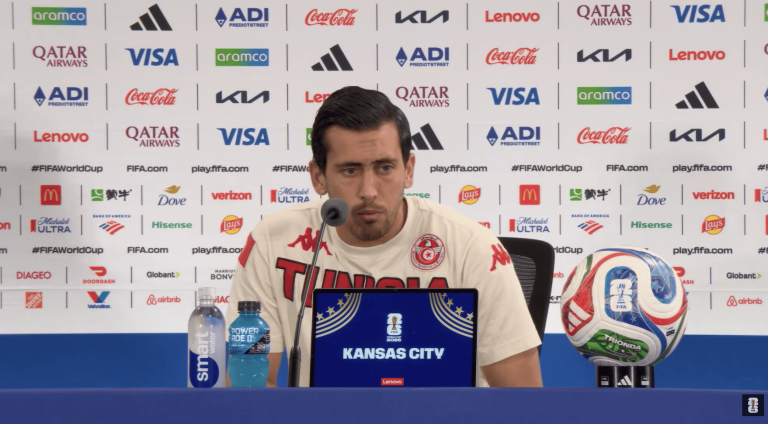 Footballer in white Tunisia shirt at a press conference, speaking into a microphone with a laptop reading 'Kansas City' and a FIFA World Cup ball on the right, sponsor backdrop behind.