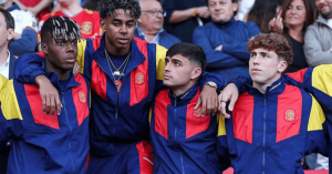 Four young men in matching blue, red, and yellow jackets with a crest, standing with arms around each other at a stadium event.