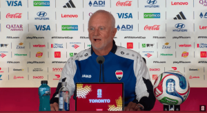 Middle-aged man in an Iraq team jersey speaks at a press conference with sponsor logos behind him; laptop on the table reads TORONTO, plus water bottles and a FIFA World Cup ball nearby.