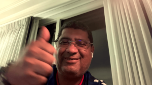 Smiling man giving a thumbs-up during a video call or selfie by a window with white curtains behind him.