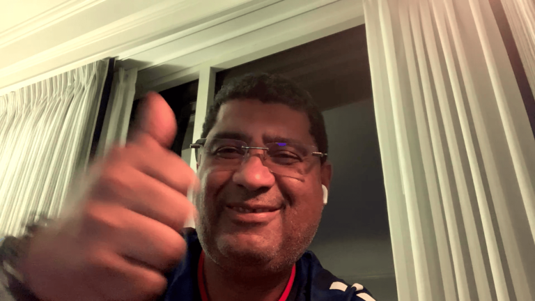 Smiling man giving a thumbs-up during a video call or selfie by a window with white curtains behind him.