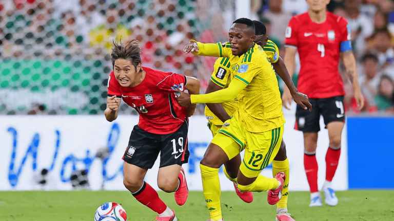 Soccer players from red and yellow teams race for the ball on a green field during a match, with a defender close behind the attacker to the left.
