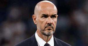 Close-up of a stern-looking man with a shaved head and gray beard in a dark suit, on a blurred stadium background.