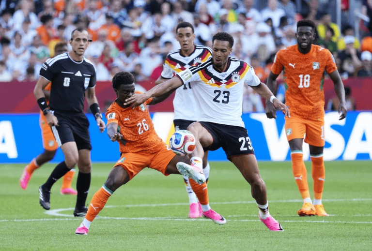 Soccer players from two teams—orange and white kits—contest a ball mid‑kick on a green field with a crowd watching.