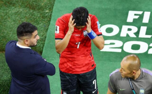 Soccer player in a red jersey number 7 covers his face with his hands on a green pitch, flanked by two staff members.