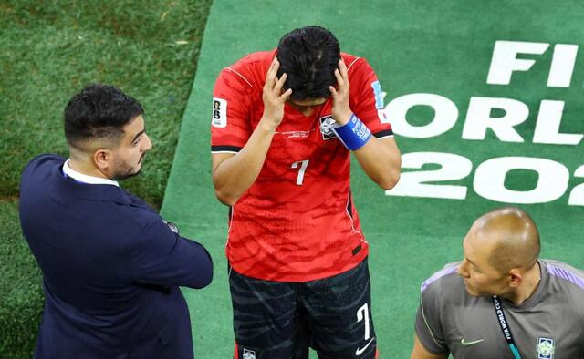 Soccer player in a red jersey number 7 covers his face with his hands on a green pitch, flanked by two staff members.