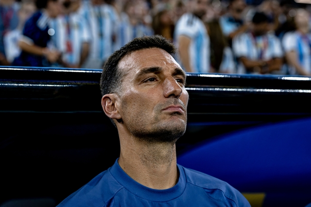 Close-up of a man in a blue jersey sitting on a bench, stadium crowd blurred in the background, looking contemplative.