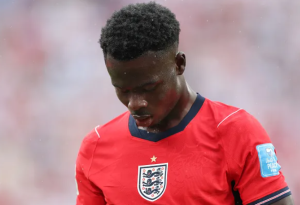 England national soccer team player in a red home kit, looking down in the rain with the crest visible on the chest.