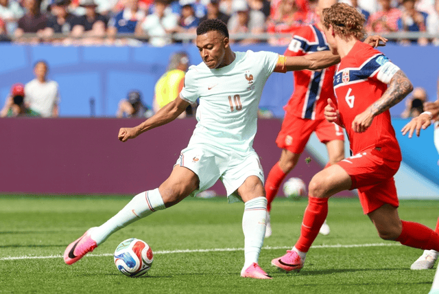 French soccer player wearing a light blue kit (#10) controls the ball while a red-jersey defender challenges him on a sunny pitch with spectators in the stands.