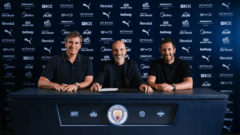Three men sit at a signing table during a press event, Manchester City backdrop behind them and the club crest on the front of the desk.