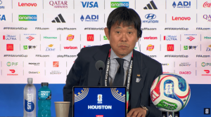 Man in a suit sits at a press conference desk with FIFA World Cup logos behind him; a soccer ball rests on the desk to his right and a laptop screen reads 'HOUSTON'.
