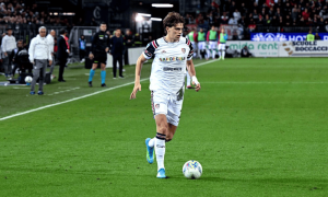 Soccer player in white uniform dribbling a ball on the green pitch with spectators in the stands behind him, during a match.