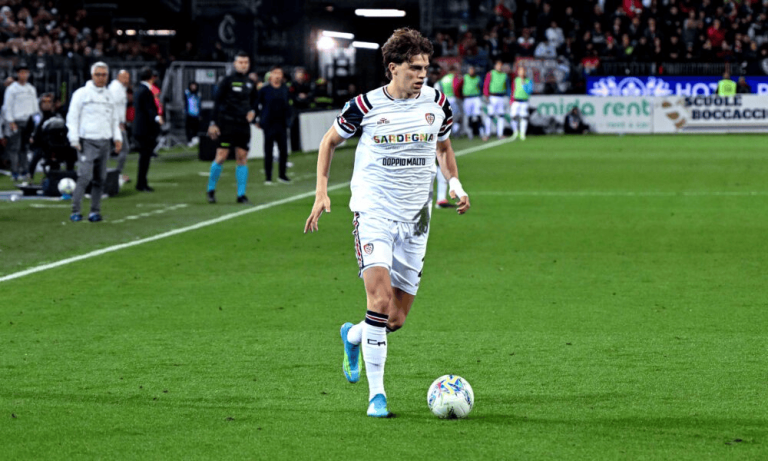 Soccer player in white uniform dribbling a ball on the green pitch with spectators in the stands behind him, during a match.