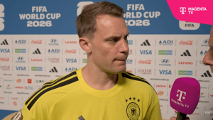 German footballer in a yellow Germany kit being interviewed, holding a Magenta TV microphone with a sponsor backdrop and 'FIFA World Cup 2026' sign.