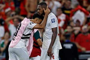 Two football players in white kits comfort each other on the pitch, one resting his head on the other's shoulder as fans look on.